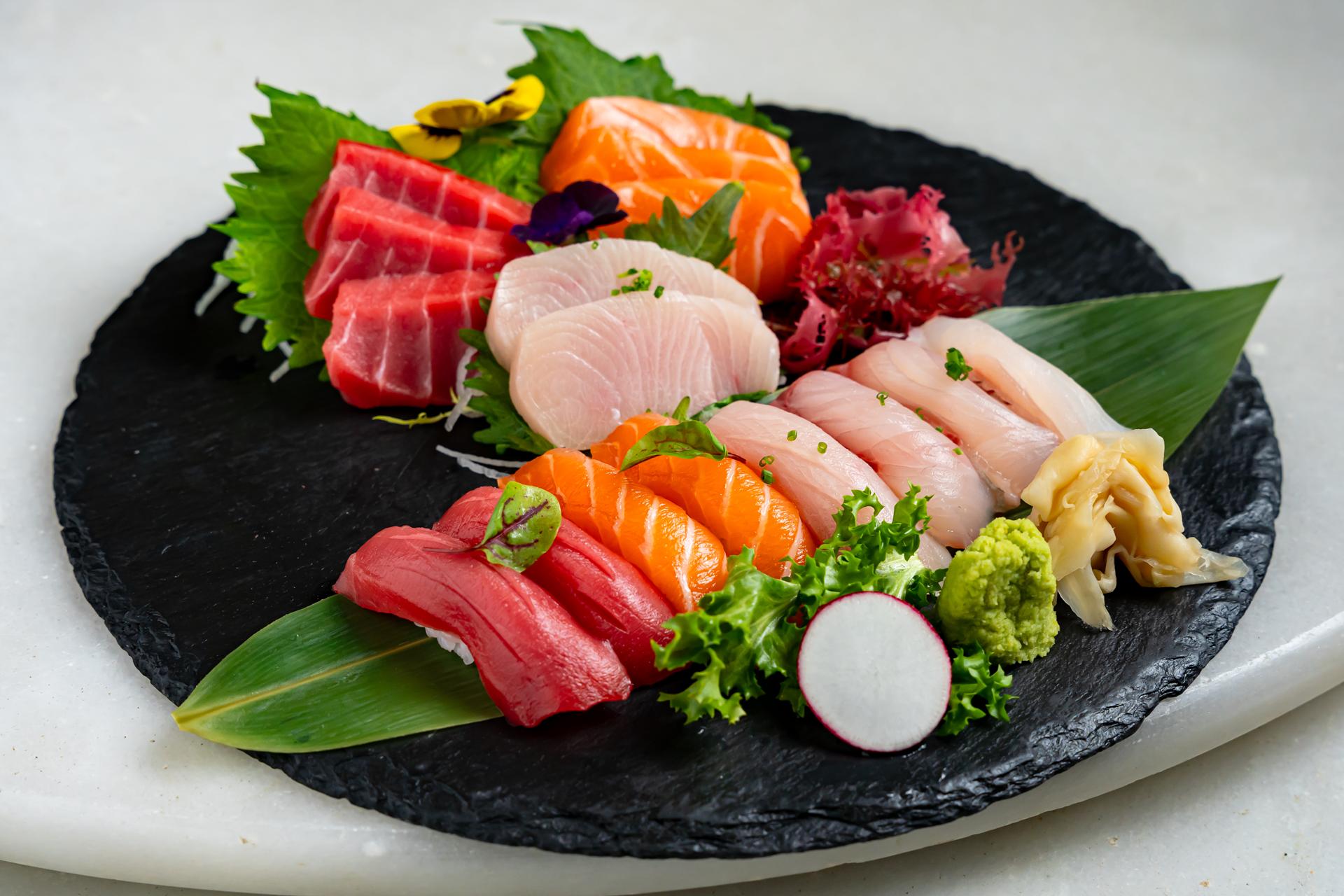 A sashimi platter of tuna, salmon and yellowtail arranged on a black slate with leaves and garnishes
