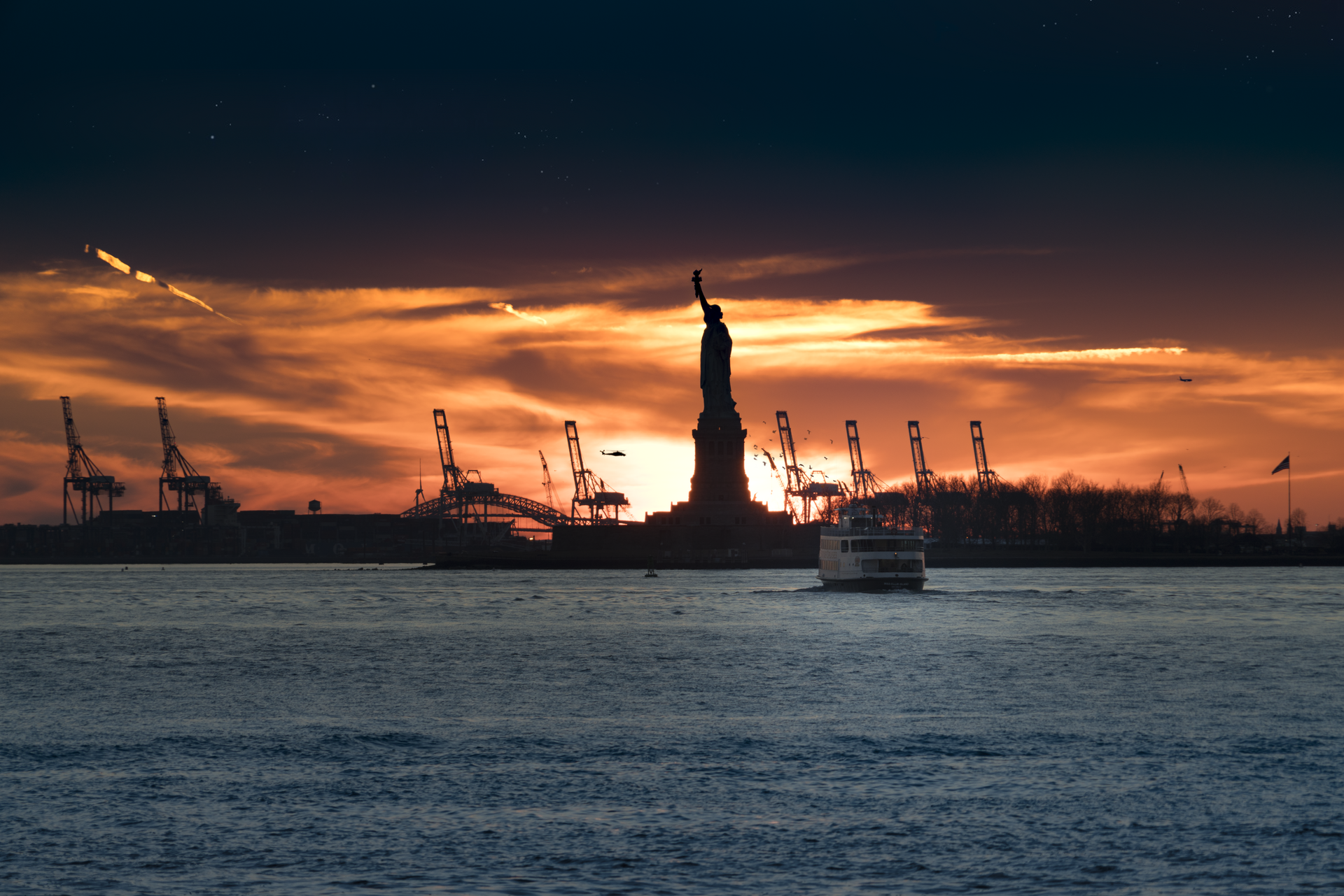 The Statue of Liberty silhouetted against a warm orange sunset, with harbor cranes on the horizon